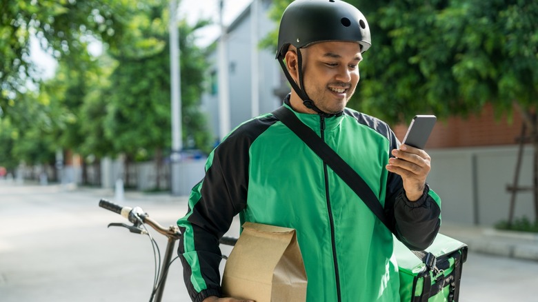 A food delivery person on a bike looks at his phone while carrying a bag