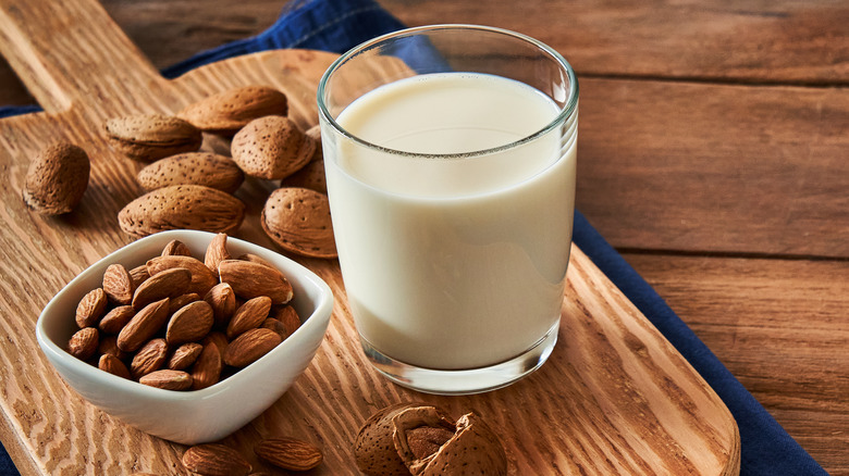Glass of almond milk on a wooden serving board and navy blue towel with a square ramekin of almonds and more almost surrounding them