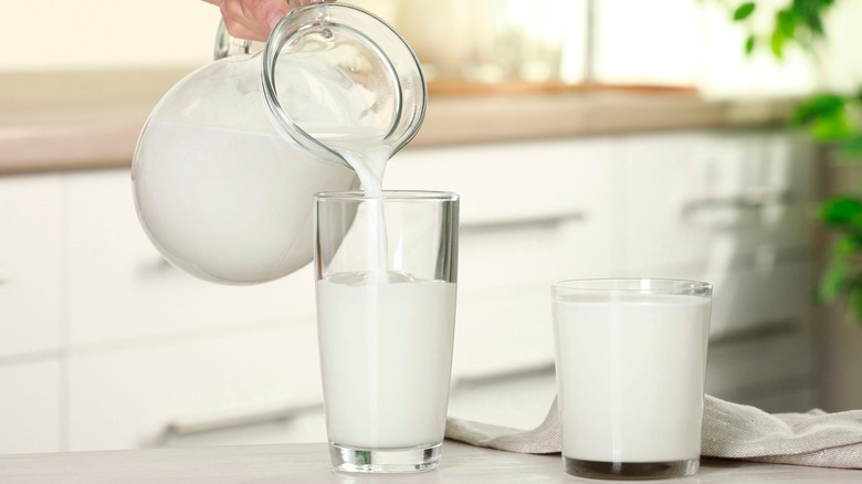 Pouring a pitcher of milk into a tall glass with a full short glass full of milk next to it in a kitchen
