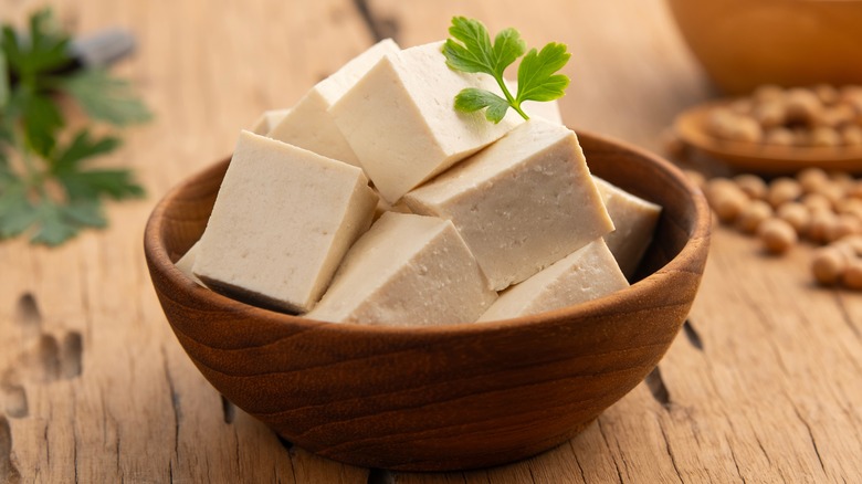 Wooden bowl of cubed raw tofu with blurred greens and soybeans in the background