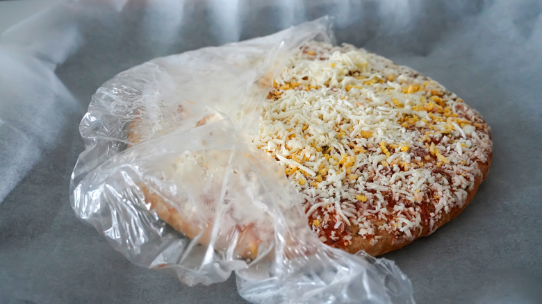 A frozen cheese pizza, with partially torn plastic packaging