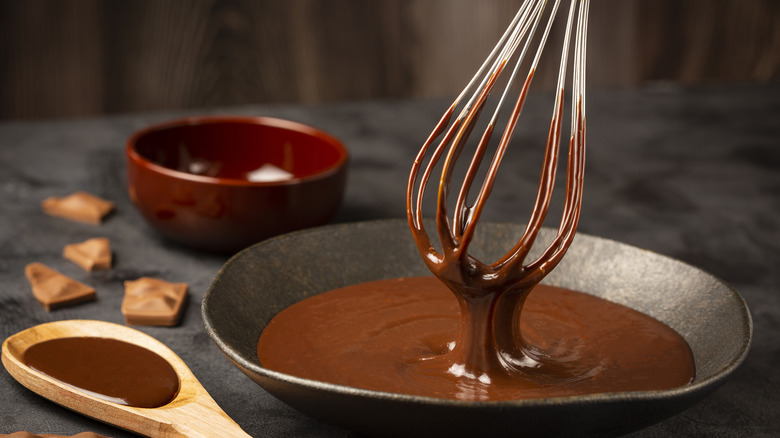Chocolate sauce in bowl with whisk on a dark background with a wooden spoon full of sauce nearby