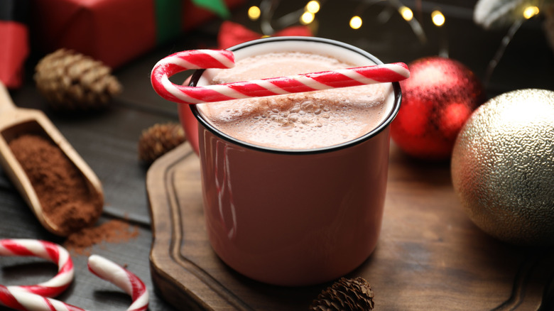 Mug of hot cocoa with a candy cane on top on a table with pinecones, ornaments, and holiday decor
