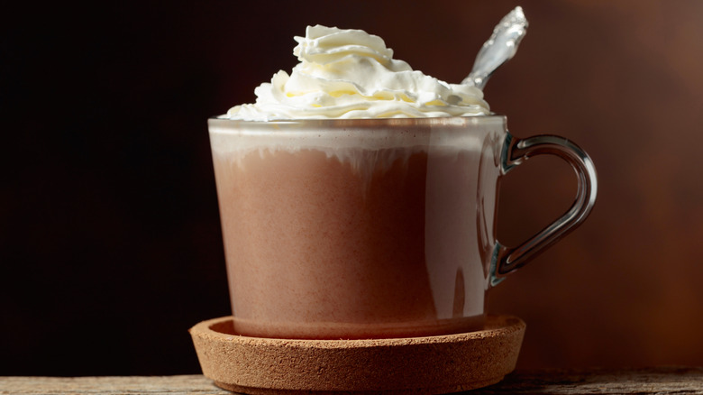 Hot chocolate with whipped cream and spoon on a cork coaster against a dark background