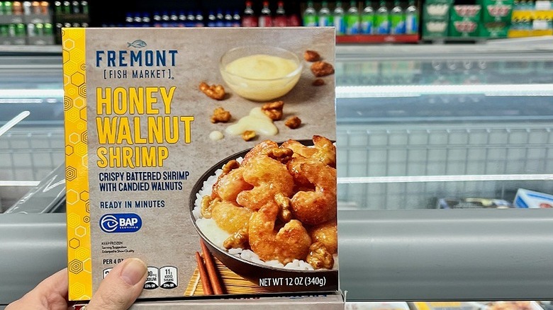 A hand holding a box of Fremont Honey Walnut Shrimp with supermarket freezer chests in the background