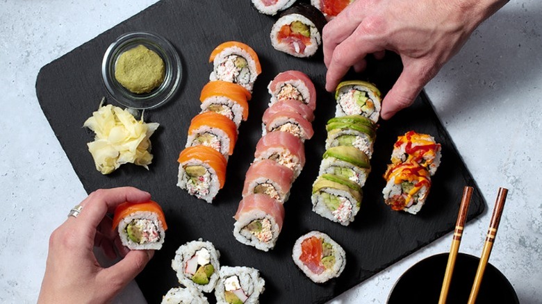 Two hands reaching for sushi rolls arranged on a black board, with wasabi and ginger on the side