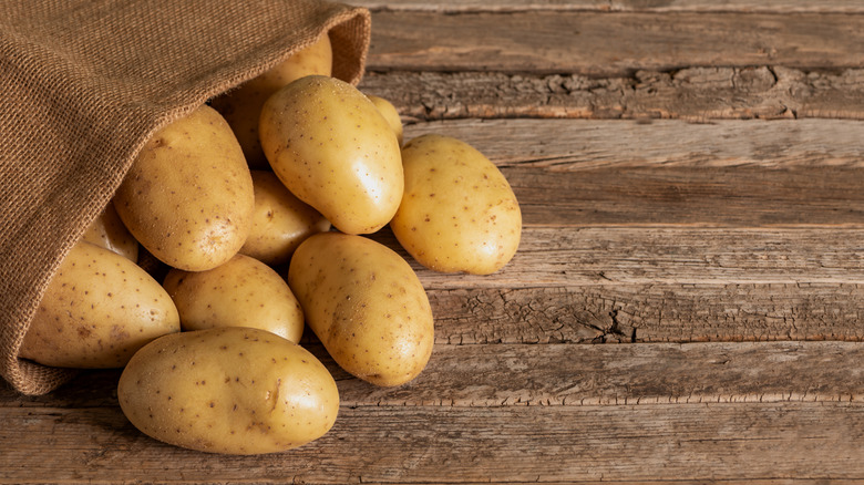 Several raw potatoes fall out of a burlap sack onto a wooden surface