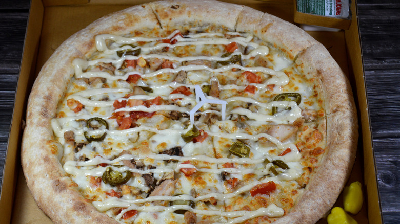 A Papa Johns pizza in a box with multiple toppings, including tomatoes, jalapeños, and a drizzled white sauce