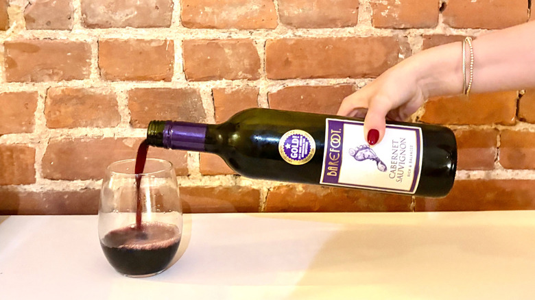 A hand reached and pours a glass of Barefoot Cabernet Sauvignon against a brick background