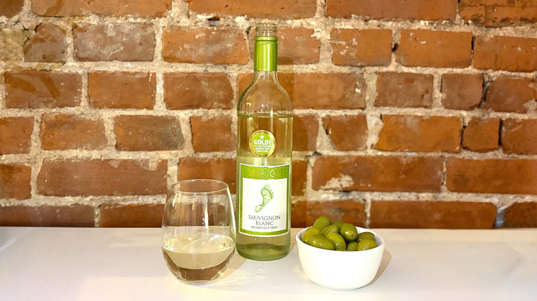 A bottle of Barefoot Sauvignon Blanc next to a glass of white wine and a bowl of olives against a brick background