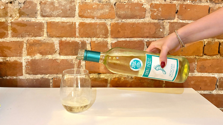 A hand reached and pours a glass of Barefoot Moscato against a brick background