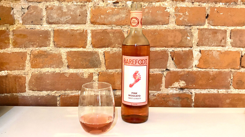 A bottle of Barefoot Pink Moscato with a poured glass next to it against a brick background