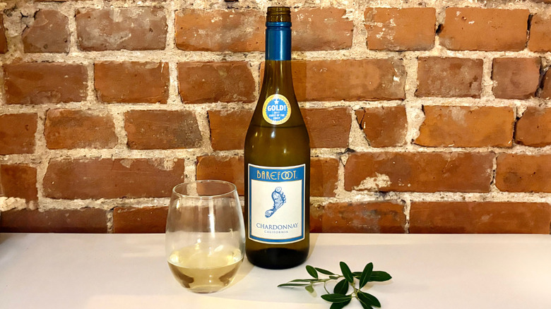 A bottle of Barefoot Chardonnay with a poured glass next to it and a green herb against a brick background