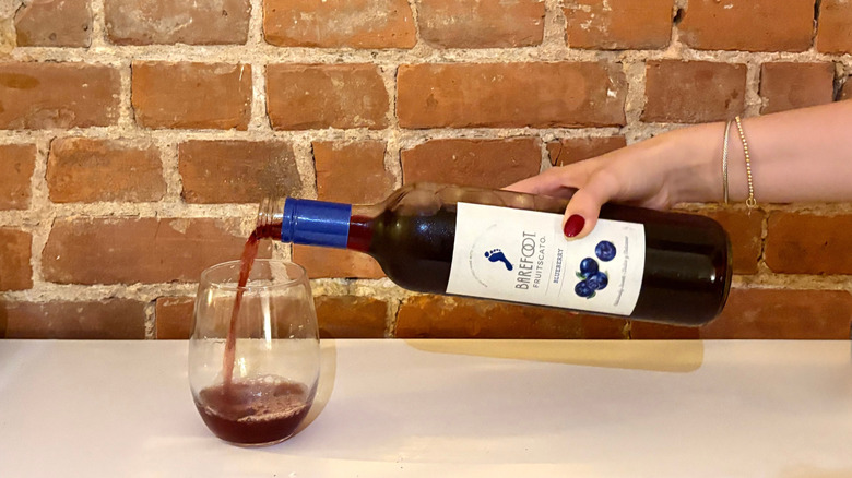 A hand reaches and pours a glass of Barefoot Blueberry Fruitscato against a brick background