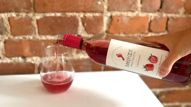 A hand reached and pours a glass of Barefoot Strawberry Fruitscato against a brick background