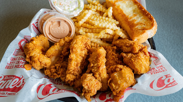 A combo meal from Raising Cane's, with its chicken fingers displayed prominently at the bottom, along with fries, Texas toast, two tubs of Cane's Sauce, and a tub of coleslaw