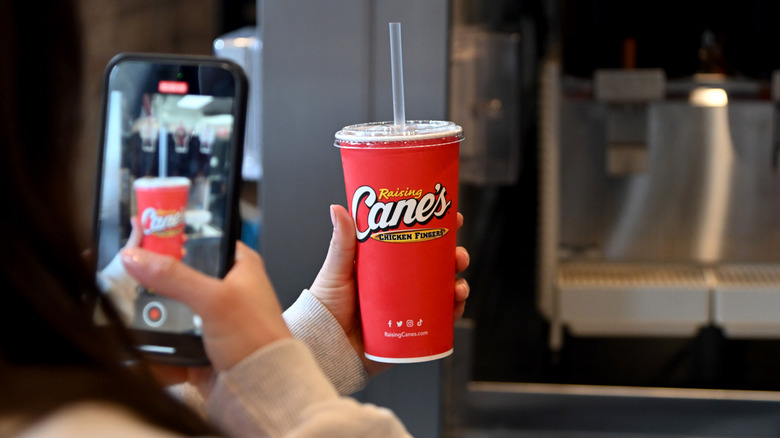 A person takes a photo of a Raising Cane's drink on their phone, while holding the drink cup in front of them