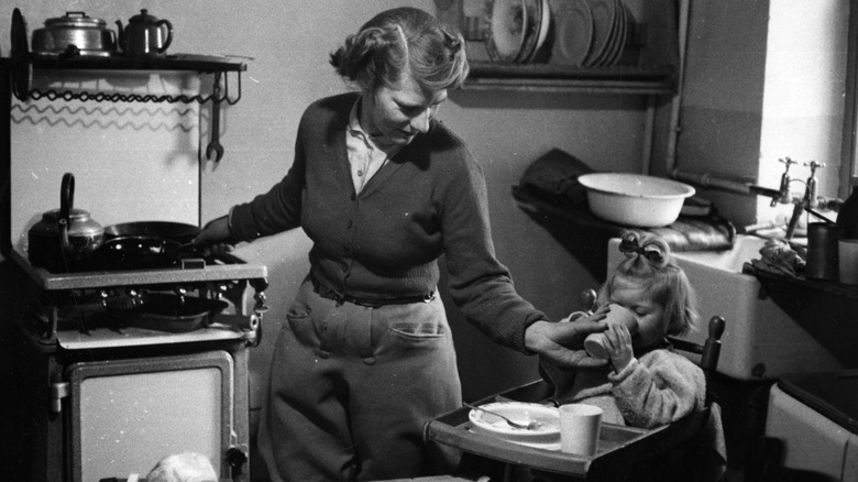 Black and white photo of a mother in the 1950s, cooking while taking care of a baby