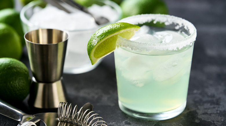 A margarita cocktail with a salt rim and a lime wedge is displayed next to cocktail equipment