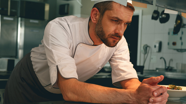 A tired, stressed chef leans forward on a counter in a restaurant kitchen