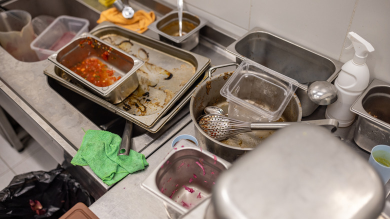 A dirty restaurant kitchen, with used utensils, trays, pans, and cloths