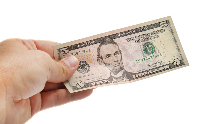 A person holds out a five dollar bill against a white background