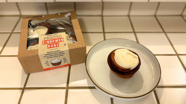 A package of Frosted Cinnamon Buns next to a plate with a frosted bun against a white tile countertop