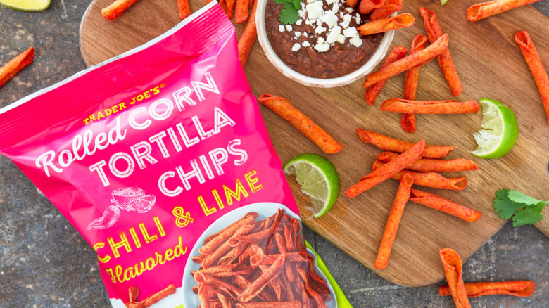 A cutting board with a bag of Trader Joe's Chili and Lime Rolled Corn Tortilla Chips surrounded by dip and lime