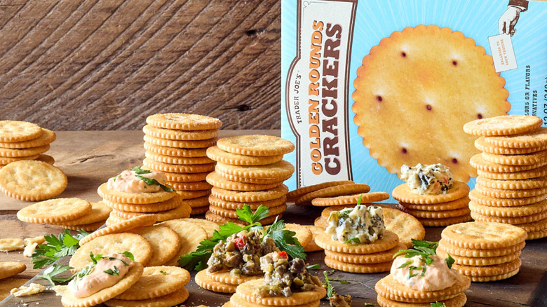 A platter of stacked Trader Joe's Golden Rounds Crackers with topping on it next to a box of the crackers as well