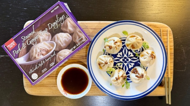 Trader Joe's Pork & Ginger Steamed Soup Dumplings on a plate next to their packaging, a bowl of soy sauce, and chopsticks