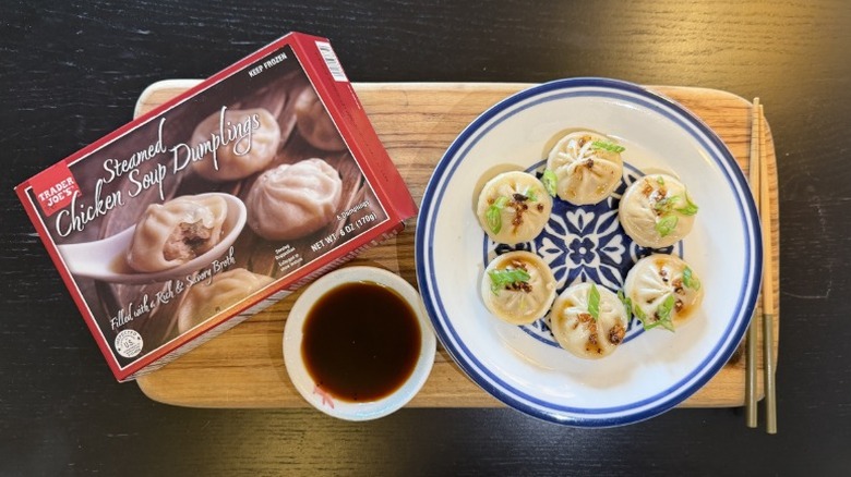 Trader Joe's Steamed Chicken Soup Dumplings on a plate next to their packaging, a bowl of soy sauce, and chopsticks