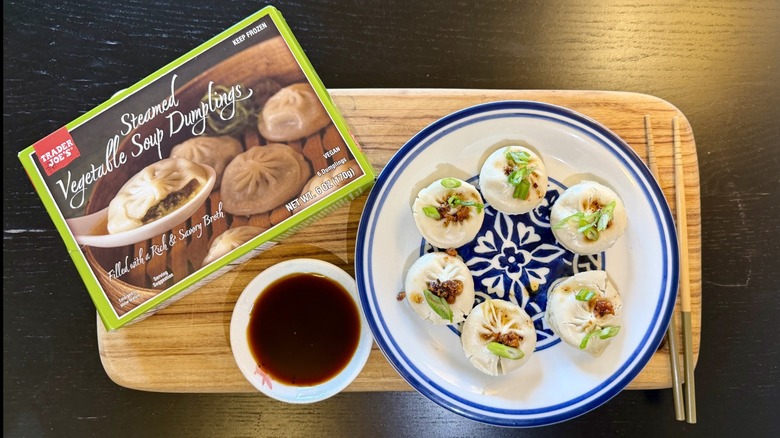 Trader Joe's Steamed Vegetable Soup Dumplings on a plate next to their packaging, a bowl of soy sauce, and chopsticks