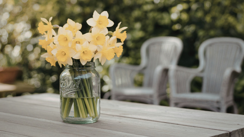 A glass jar holding bright yellow flowers and sitting on top of a wooden outdoor table