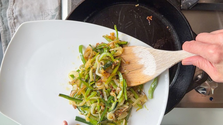 Transferring caramelized sliced poblano peppers and sweet onions to a plate from a cast iron skillet on stovetop with wooden spoon