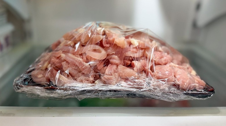 Sliced raw chicken thighs piled on plate and covered with plastic in refrigerator