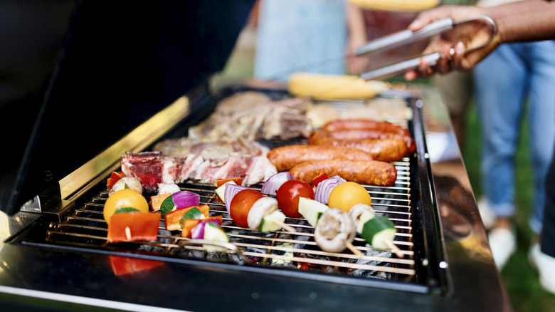 A hand holding tongs over a gas grill filled with cooking food