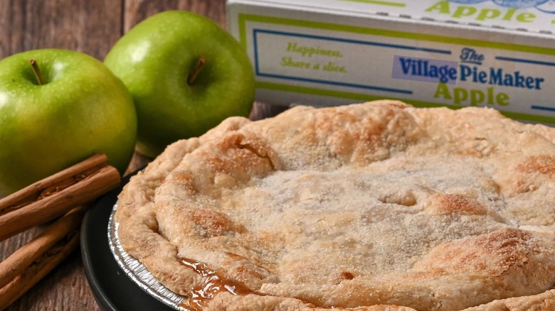 Close up of an apple pie from The Village PieMaker next to green apples and cinnamon sticks with a box in the background