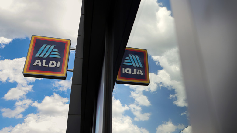 An Aldi store sign against a cloudy blue sky, reflected in the store's window