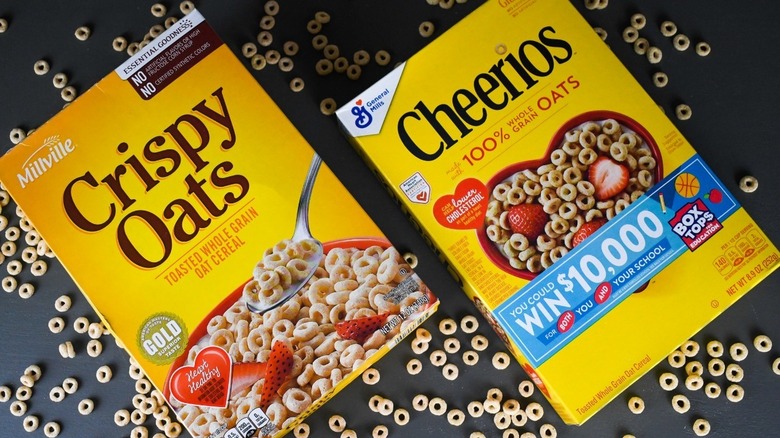 Side by side of Millville crispy oats cereal and General Mills Cheerios cereal boxes with loose cereal in the background