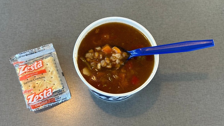 Cup of Vegetable Beef Soup from Culver's with a blue plastic spoon in it accompanied by a package of Zesta saltine crackers