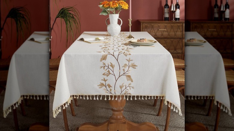 White tablecloth with leaf design sits on table with flowers, a candle, and a plate with food