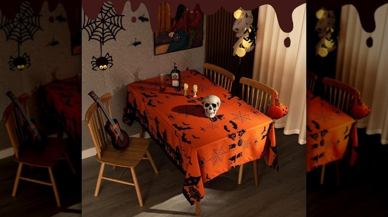 Overview of orange and black Halloween tablecloth on a table with candles, and bottle, and a skull decoration in a Halloween-decorated room