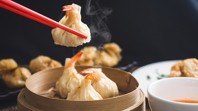 Red chopsticks lifting shrimp dumplings out of bamboo steam basket on dim sum platter.