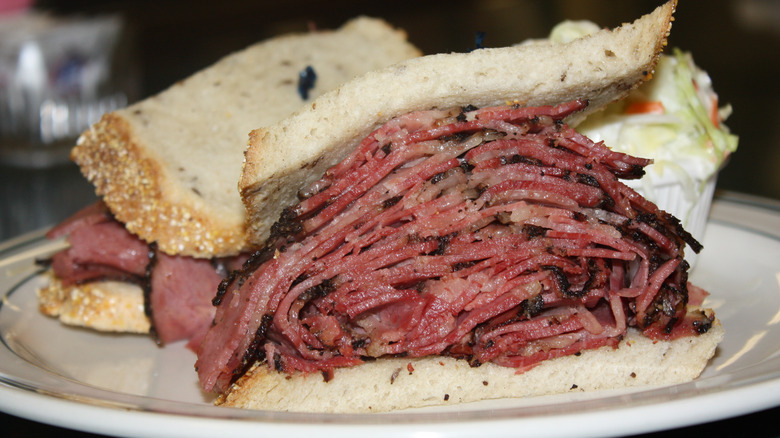 A closeup of a pastrami sandwich cut in half with a side of cole slaw on a white plate
