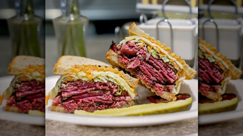 A pastrami sandwich with cole slaw cut in half and served with a pickle spear on a white plate