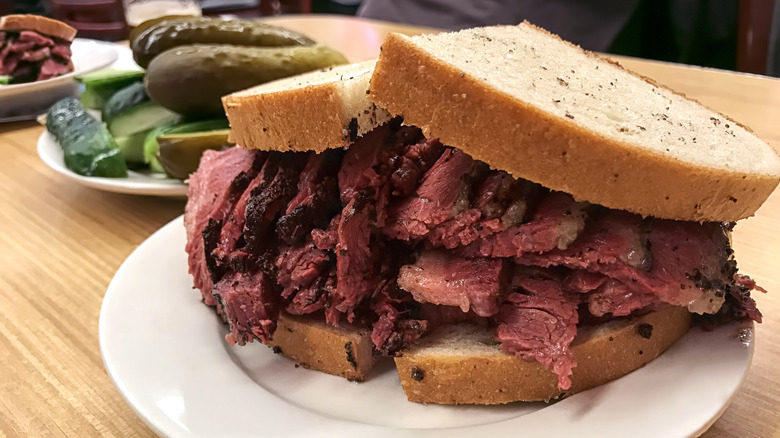 A sandwich with pastrami piled high next to a plate of pickles on a table