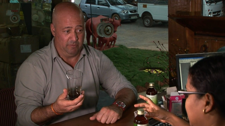 Andrew Zimmern holding a tall glass with a small amount of dark brown liquid in it