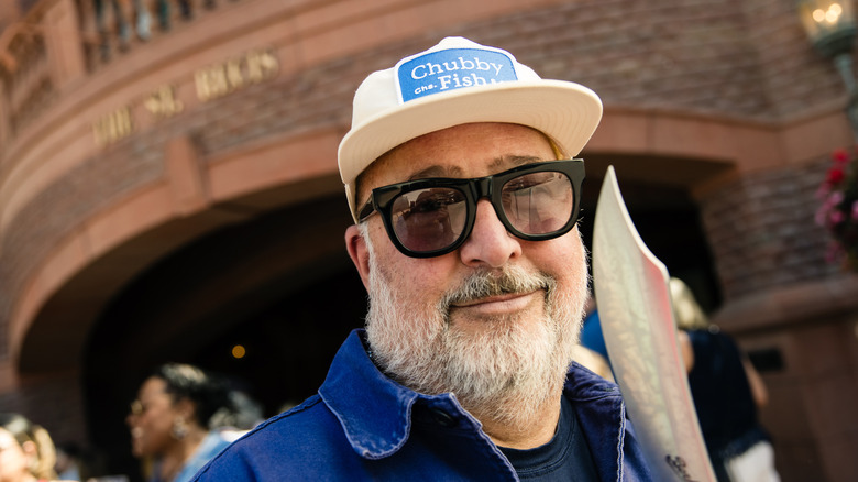 Andrew Zimmern in front of a building in a hat and sunglasses