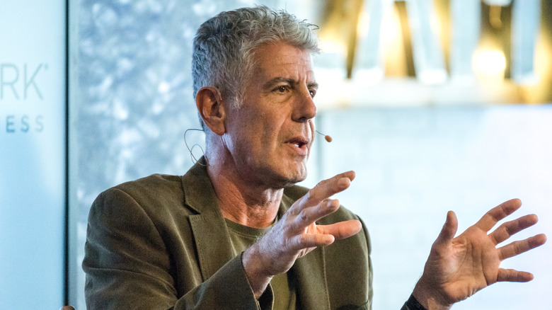 Anthony Bourdain speaking on stage in a green suit jacket and t-shirt, talking with his hands