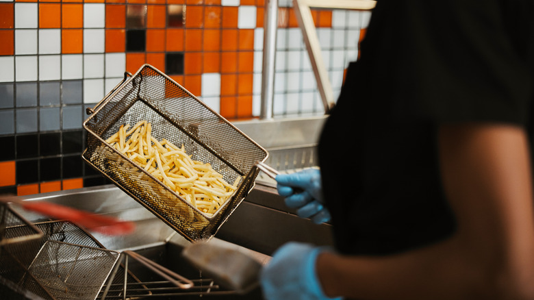 Fast food worker with gloves on holding basket of fries headed for deep fryer.